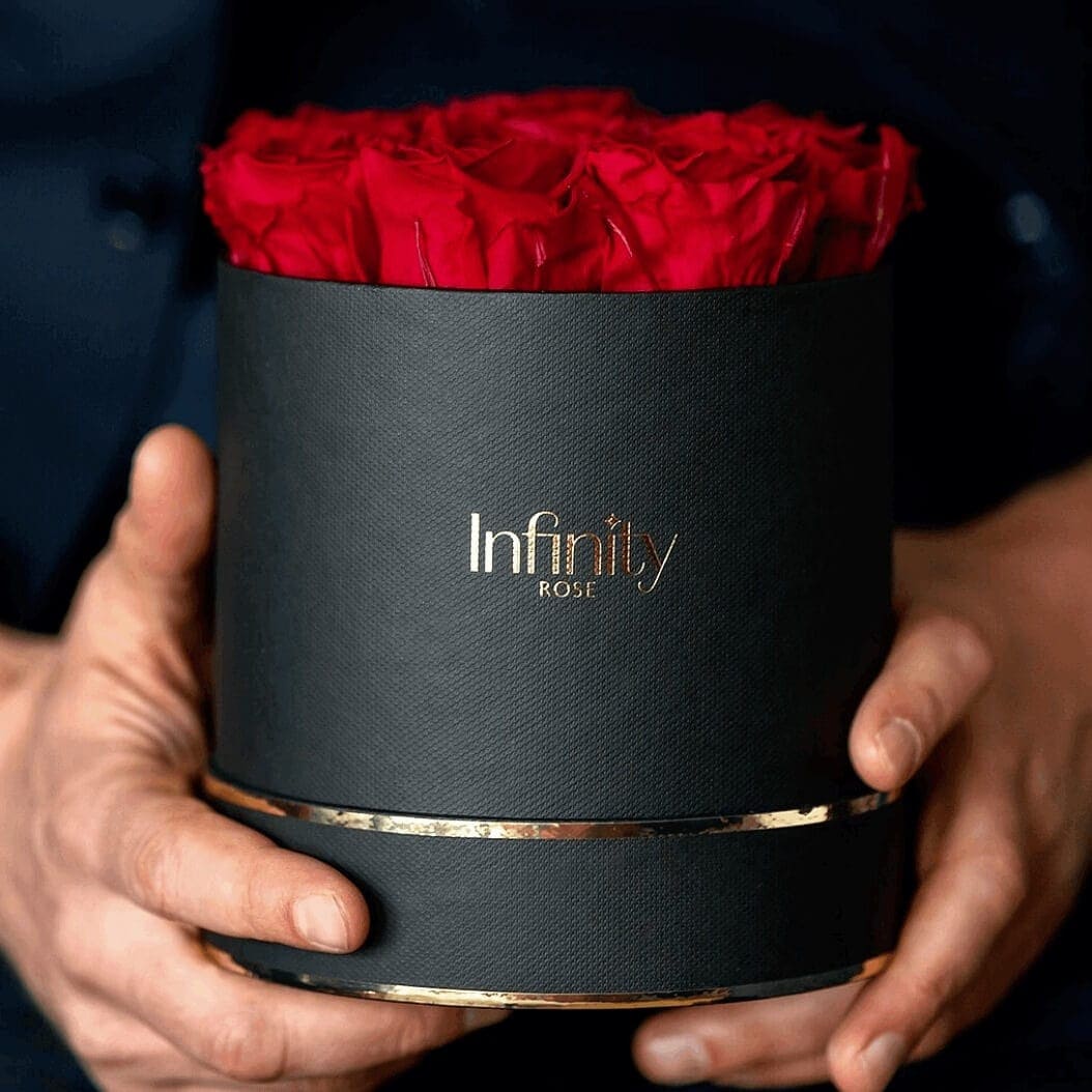 Wieczne róże flower box firmy Infinity Rose trzymane w dłoniach podarowane przez mężczyznę jako prezent dla kobiety na Walentynki i Dzień Kobiet czerwone kwiaty i czarne złote luksusowe pudełko